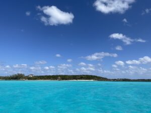 crystal clear turqoise waters are the sea, with a tropical beach with green foliage and a big blue sky overhead with some white fluffy clouds