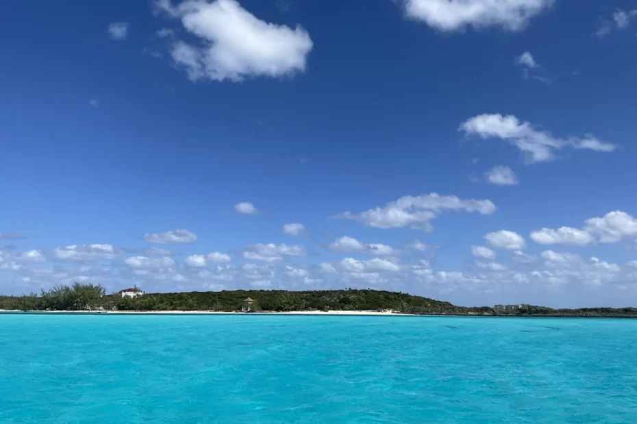 crystal clear turqoise waters are the sea, with a tropical beach with green foliage and a big blue sky overhead with some white fluffy clouds