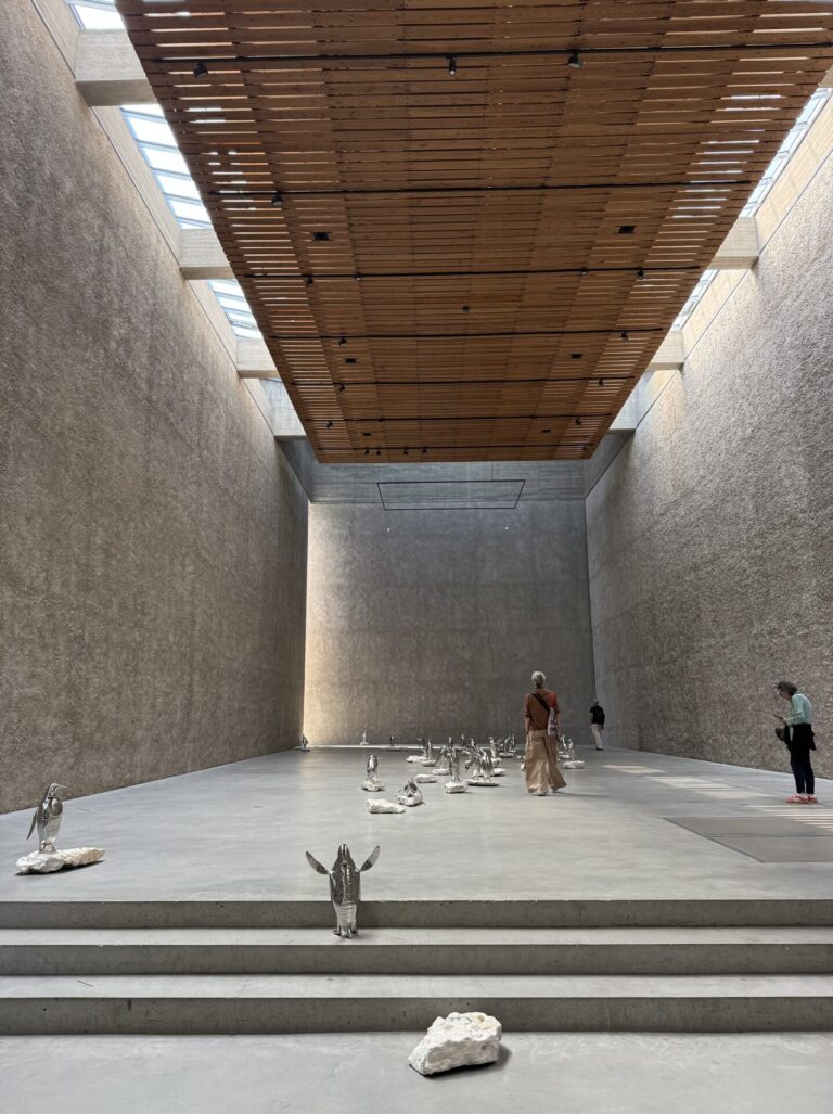 A long, tall room with concrete walls and a wooden ceiling structure. Along the floor are chrome penguins in different positions.